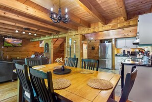 A wooden interior with a dining table, chairs, a chandelier, and a kitchen area.