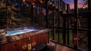A hot tub with a wooden exterior, a bottle of wine, and glasses on a bench, set against a backdrop of a starry night sky visible through large windows.