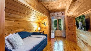 A wooden cabin room with a bed, a ceiling fan, a window with curtains, and a wall-mounted TV.