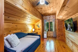 Quarto de cabana de madeira com cama, ventilador de teto, janela com cortinas e televisão na parede.