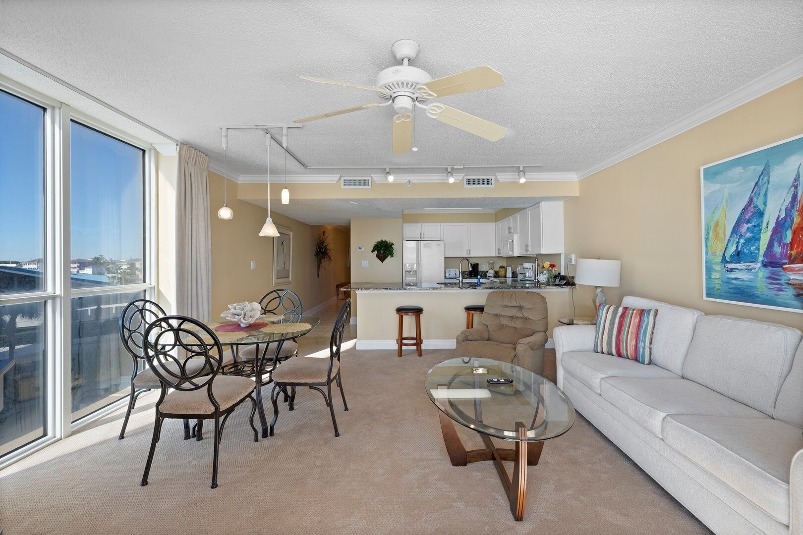 A modern living room with a glass coffee table, a sofa, and a dining area with chairs. There is a kitchen in the background with white cabinets and a ceiling fan.