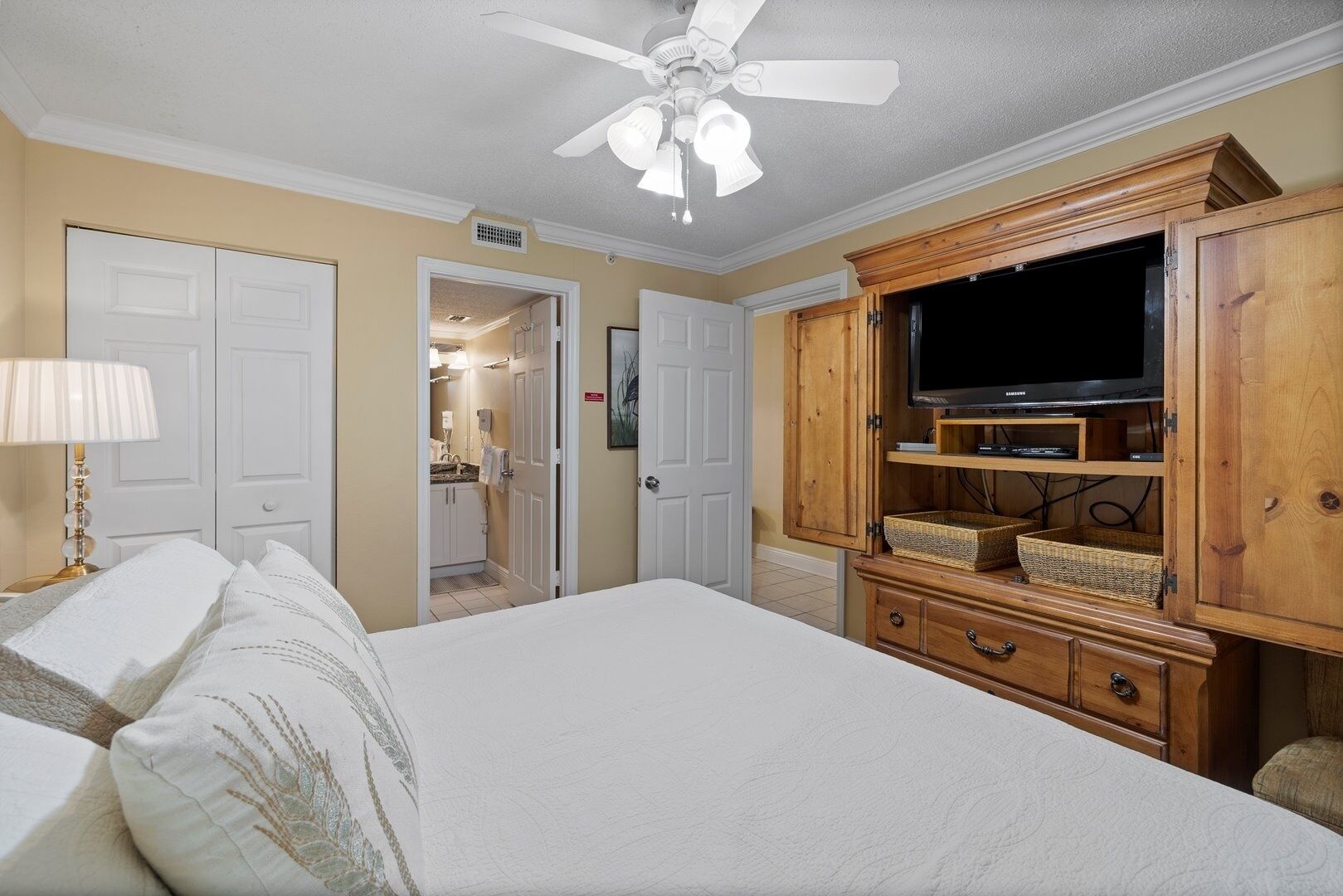 A bedroom with a bed, a ceiling fan, a television on a wooden cabinet, and a bathroom visible through an open door.