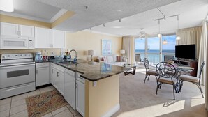 A modern kitchen with white cabinets, a granite countertop, and a dining area with a round table and chairs. - Pelican Beach 0411 1 Bedroom Condo by Pelican Beach Management (Destin)