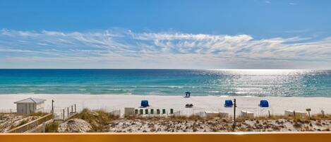 A beach view from a balcony with a wooden railing, featuring a sandy beach, a small structure, and several blue beach chairs.