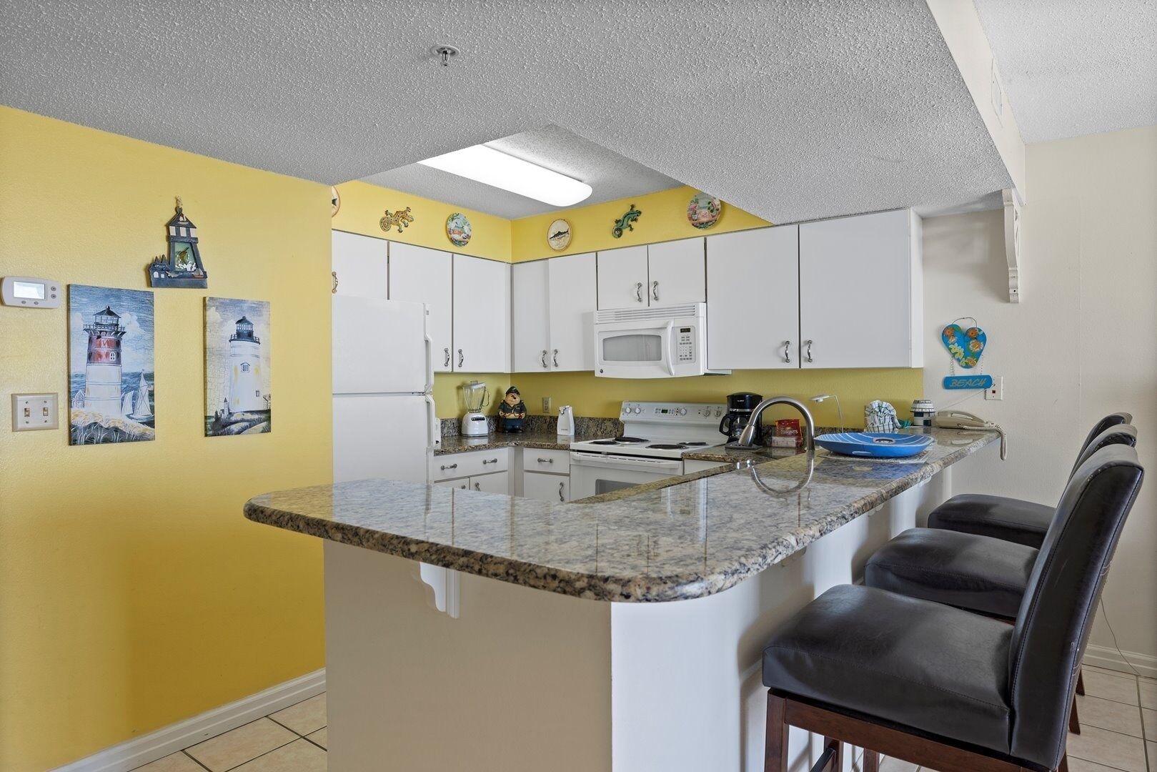 A compact kitchen with white cabinets, a microwave, a stove, and a granite countertop. There are two chairs and a small island with a sink.