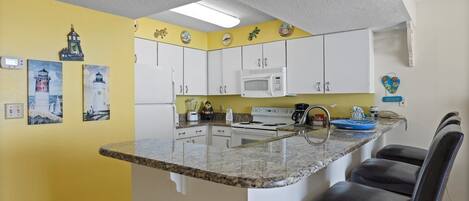 A compact kitchen with white cabinets, a microwave, a stove, and a granite countertop. There are two chairs and a small island with a sink.