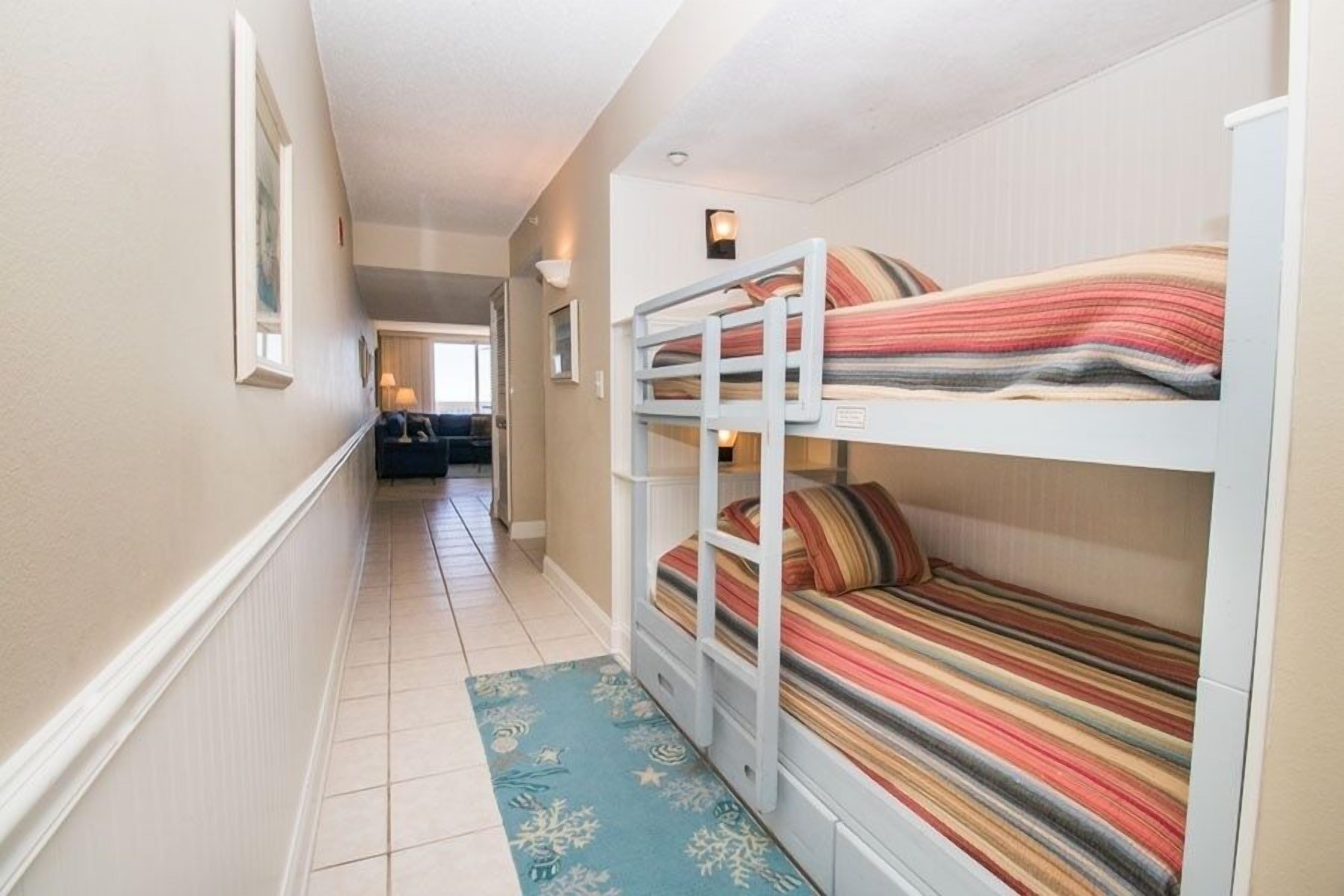 A narrow hallway with bunk beds on the right, a door at the end, and a blue patterned rug on the floor.