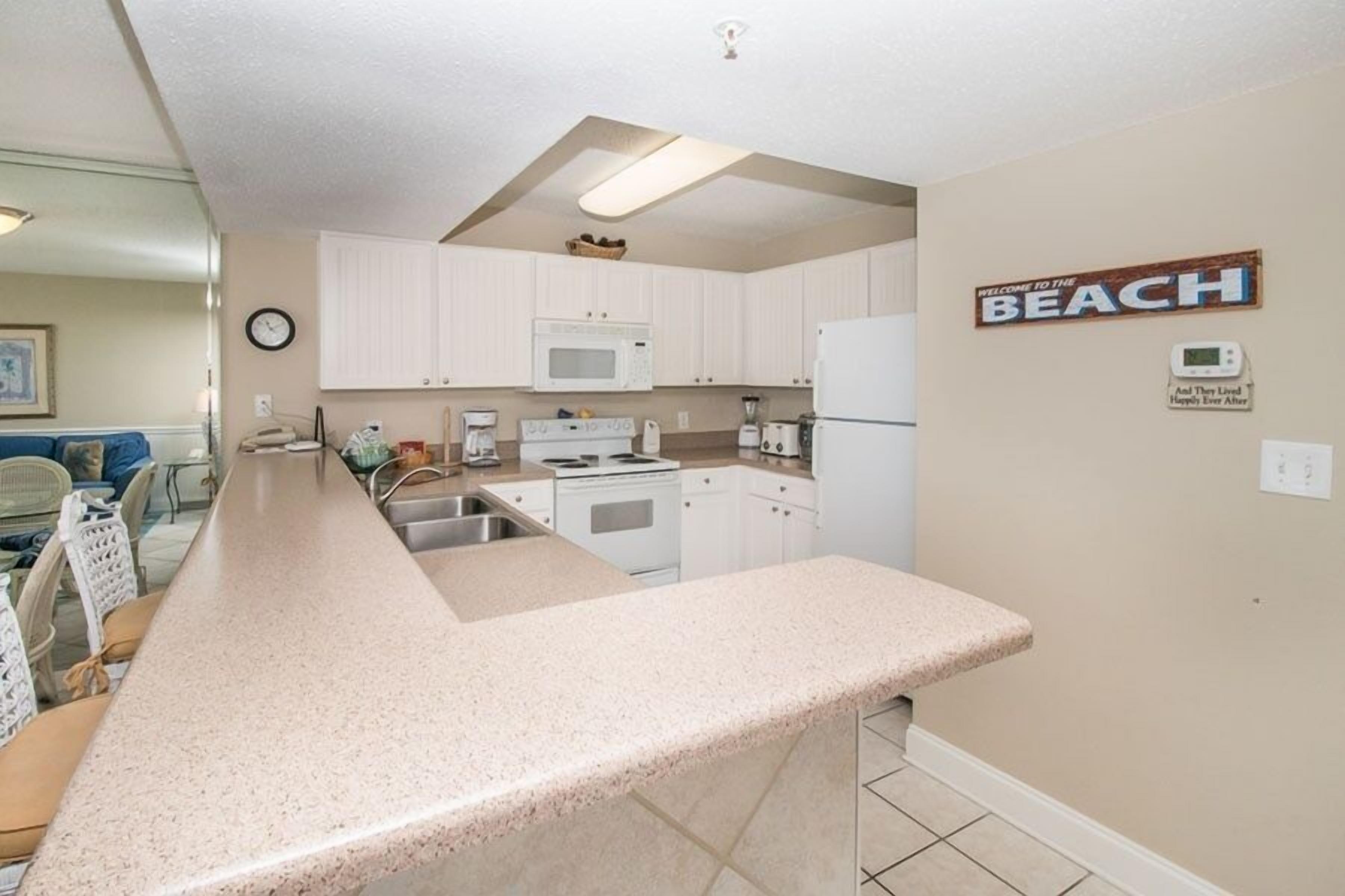 A kitchen with white cabinets, a microwave, stove, and a refrigerator. There is a sign on the wall that says "Welcome to the Beach".