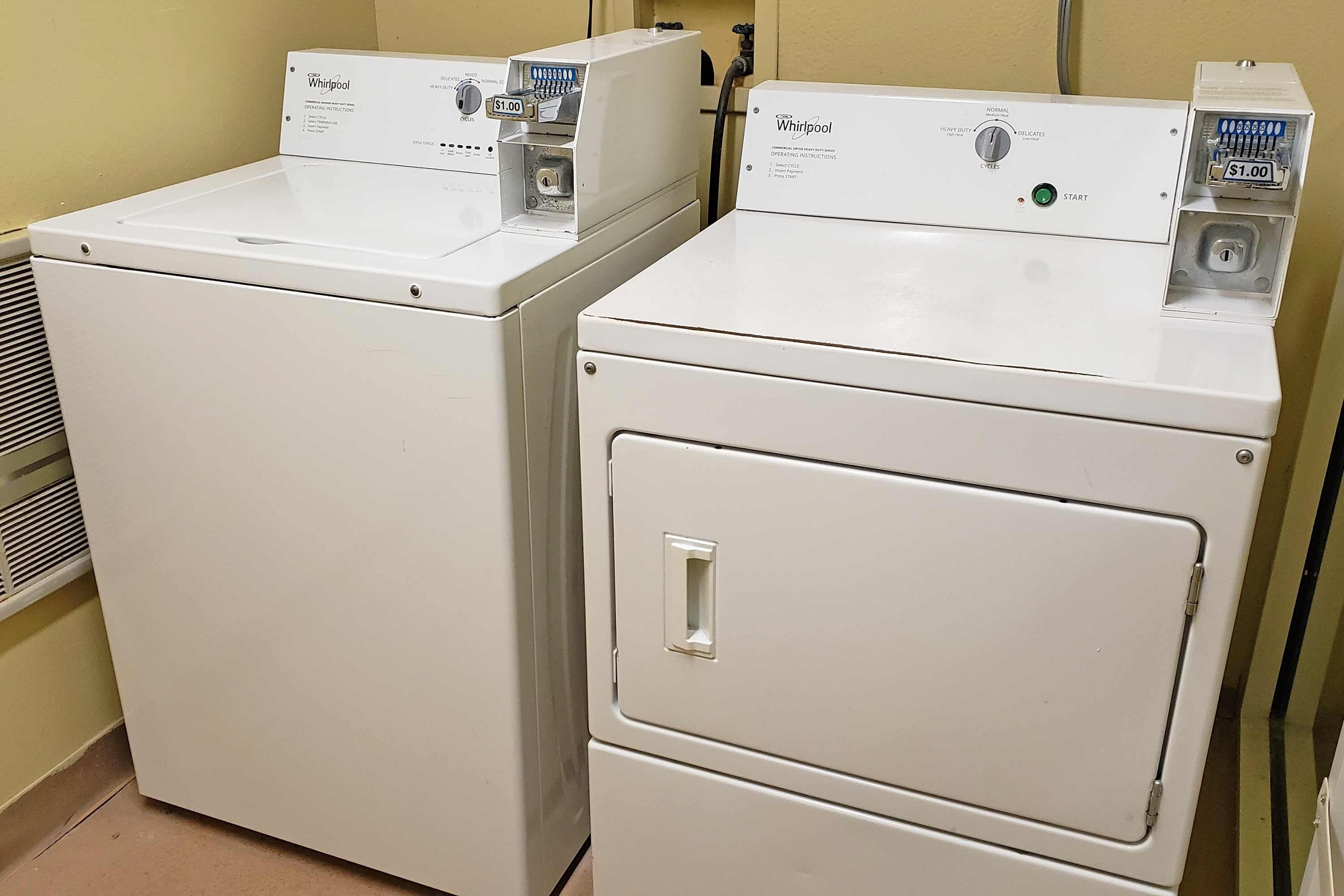 Two white Whirlpool front-loading washing machines in a laundry room with beige walls and a vent on the left.