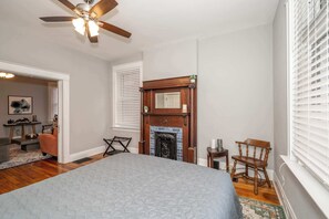 A bedroom with a large bed, a wooden fireplace, a ceiling fan, and a window with blinds. - Suites on Main 1 (Covington)
