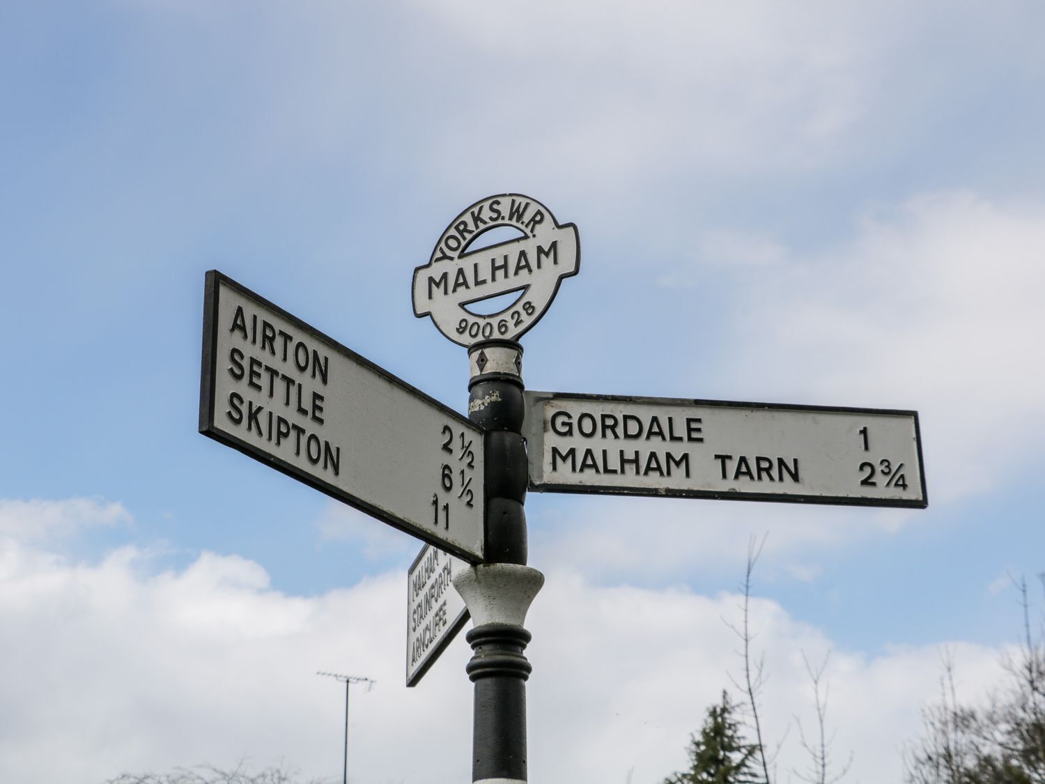 A directional signpost with multiple arms pointing to various destinations such as Airton, Settle, Skipton, Gorda, Malham Tarn, and Malham.