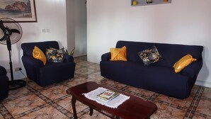 Ein Wohnzimmer mit zwei dunkelblauen Sofas, jedes mit einem gelben Kissen, einem Holztisch mit einer Zeitschrift und einem Standventilator.