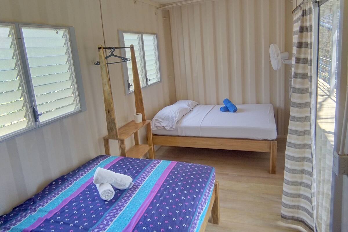 A small, single-room accommodation with a bed, a small table, and a window with blinds.