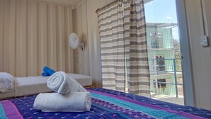 A room with a bed, a small sofa, a window with curtains, a fan, and a view of a building outside. - Casa Maya Eco Lodge (San Juan del Sur)