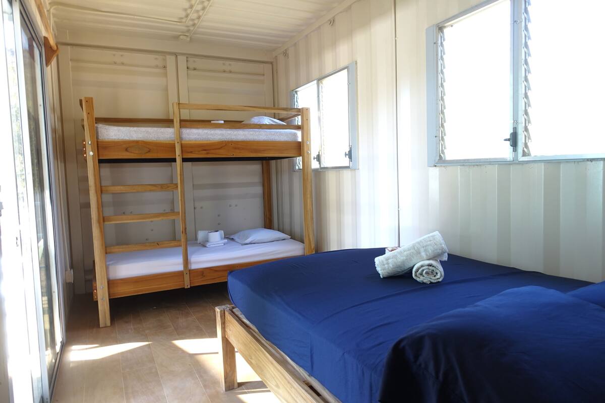 A room with a bunk bed and a single bed, both with white bedding. There is a window with blinds, a door, and a towel on the single bed.