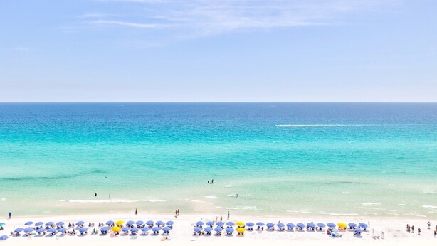 A beach with clear turquoise water, white sand, and numerous umbrellas. - Pelican Beach 1206 1 Bedroom Condo by Pelican Beach Management (Destin)