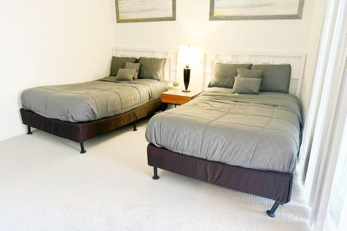 A room with two beds, a nightstand with a lamp, and artwork on the wall.