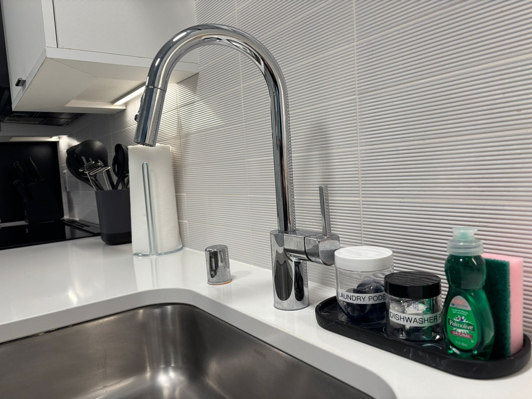A modern kitchen with a stainless steel sink, a faucet, and a tray holding various items like a dishwashing liquid bottle, a container labeled "laundry pods," and a small cup.