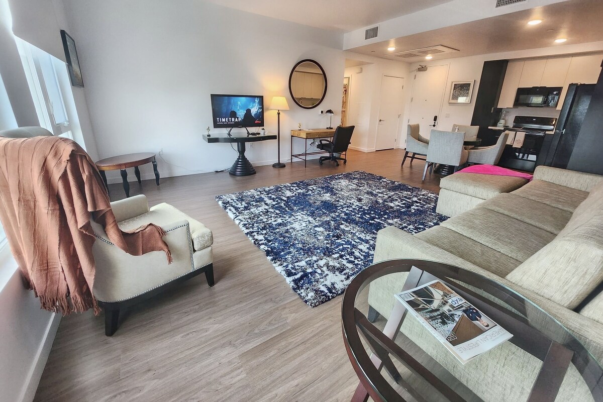 A modern living room with a sofa, armchair, and a coffee table with magazines. There is a TV, a desk with a chair, and a kitchen area in the background.