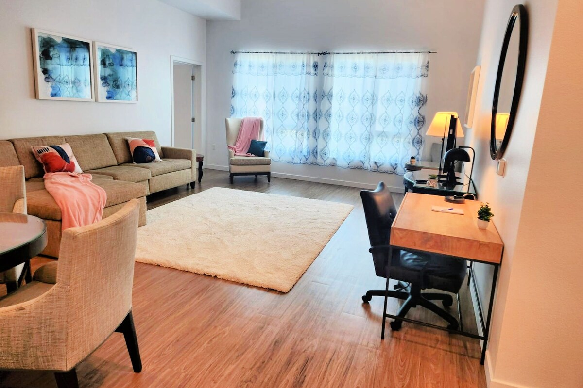 A modern living room with a sofa, armchair, and a desk with a chair. There are two framed artworks on the wall, a window with patterned curtains, and a door in the background.