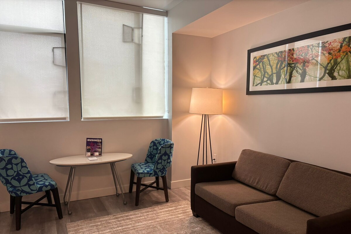 A room with a brown sofa, two patterned chairs, a small table, a floor lamp, and a framed picture on the wall.