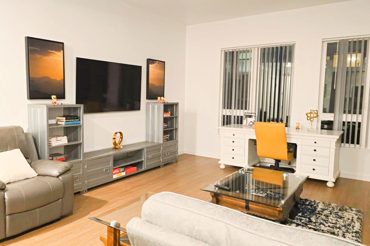 A modern living room with a grey sofa, a glass coffee table, and a white armchair. There are two grey bookshelves with books and decorative items. A large window with vertical blinds is on the right.