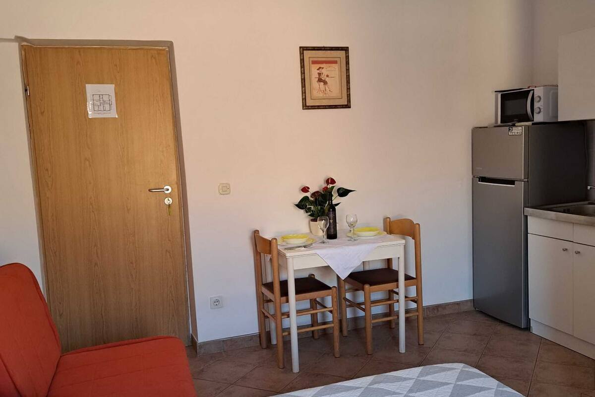 A small, compact room with a wooden door, a dining table with chairs, a small kitchen area with a refrigerator and microwave, and a bed with a patterned bedspread.