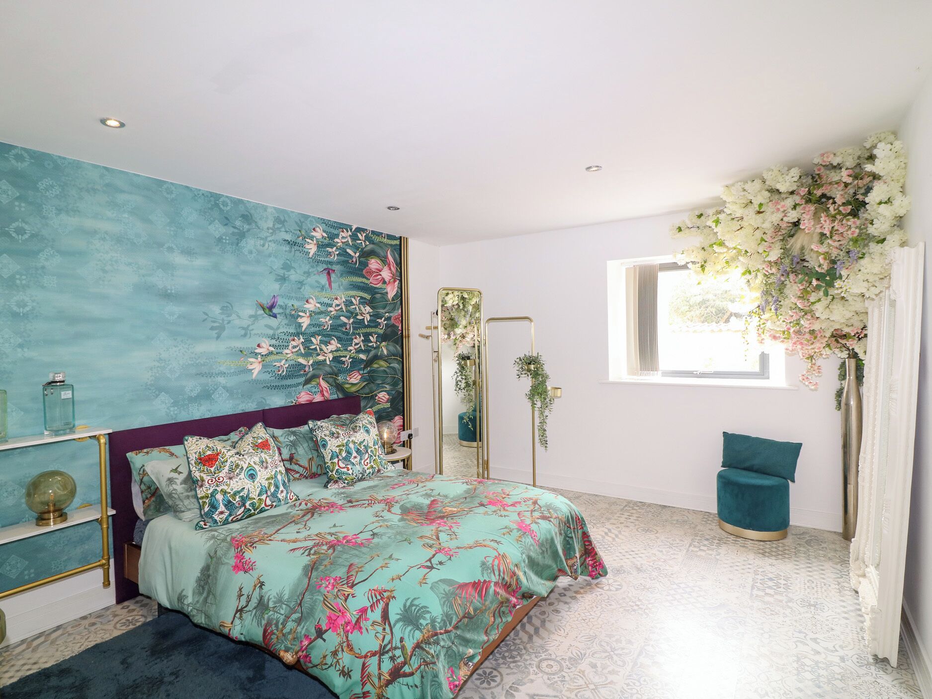 Ein Schlafzimmer mit einem Bett, einem Spiegel, einem Fenster mit Blumen und einem blauen Hocker.