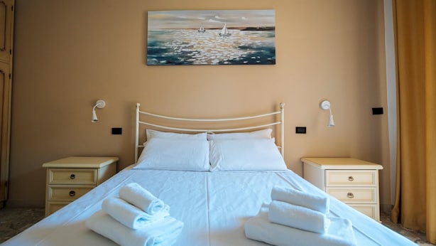A neatly made bed with white linens and towels, a painting of sailboats on the wall, and two bedside tables with lamps.