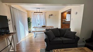 Ein Wohnzimmer mit einem dunklen Sofa, einem Couchtisch aus Holz und einem Fernseher auf einem Lowboard. Im Hintergrund ist eine Essecke mit Tisch und Stühlen zu sehen.