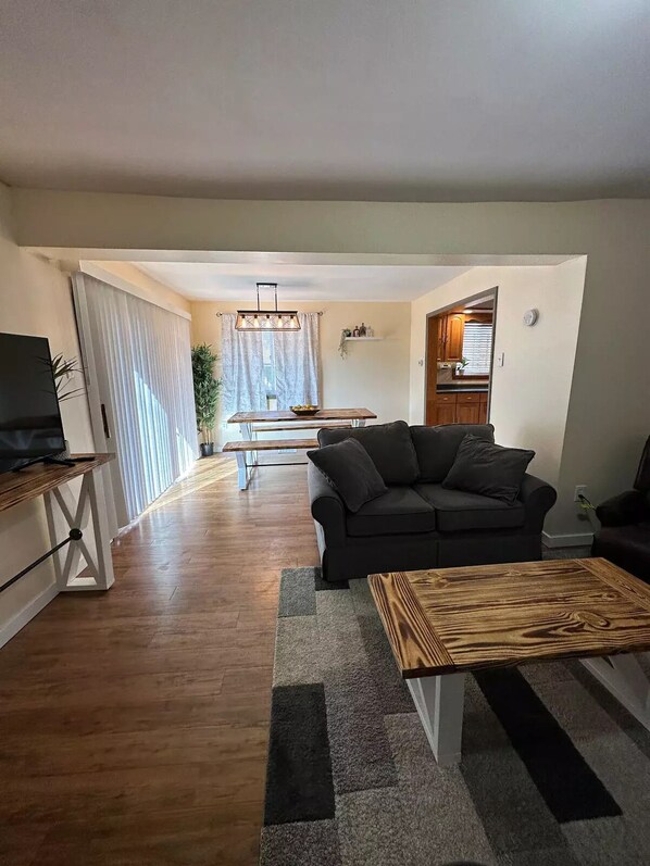 Uma sala de estar com um sofá escuro, uma mesa de centro de madeira e uma TV em um móvel. Ao fundo, há uma área de jantar com mesa e cadeiras.