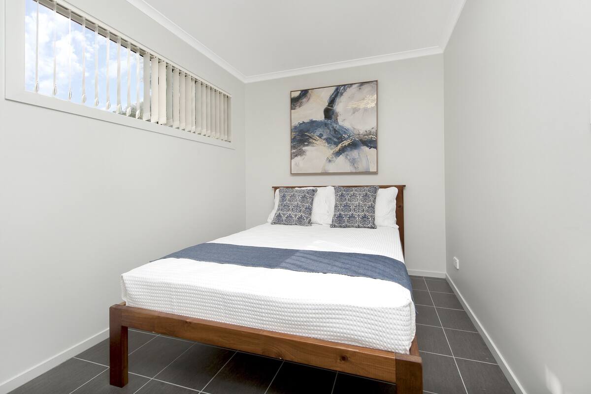 A bedroom with a bed, a window with blinds, a painting on the wall, and a tiled floor.