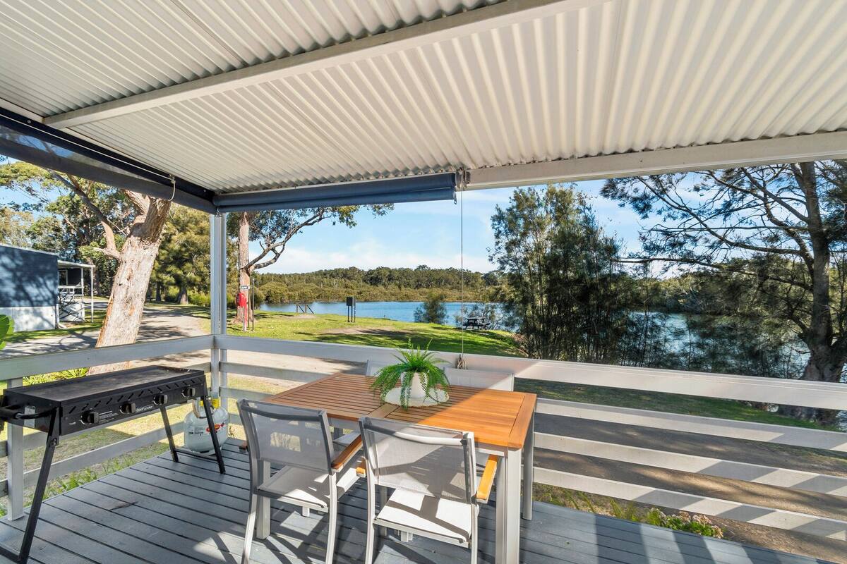 A covered outdoor area with a wooden table and chairs, overlooking a lake and trees.