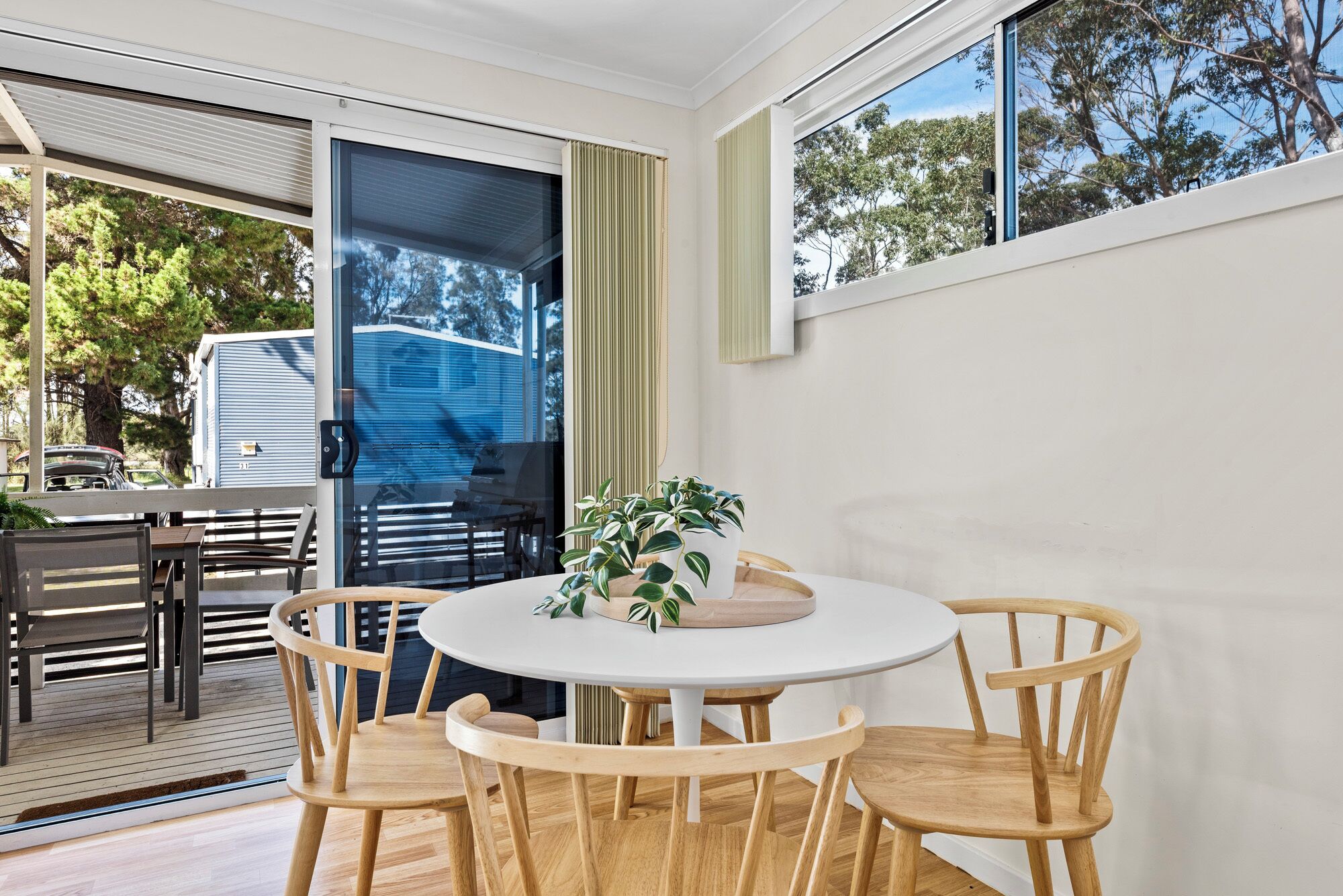 Un comedor moderno con una mesa redonda y sillas de madera, ventanas grandes con cortinas, y vistas a árboles y un vehículo afuera.