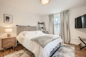 Quarto com cama, mesa de cabeceira, abajur, televisão e janela com cortinas.
