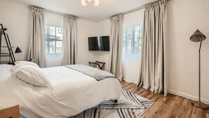 Une chambre à coucher avec un lit, un téléviseur à écran plat, une lampe sur pied et une fenêtre avec des rideaux.
