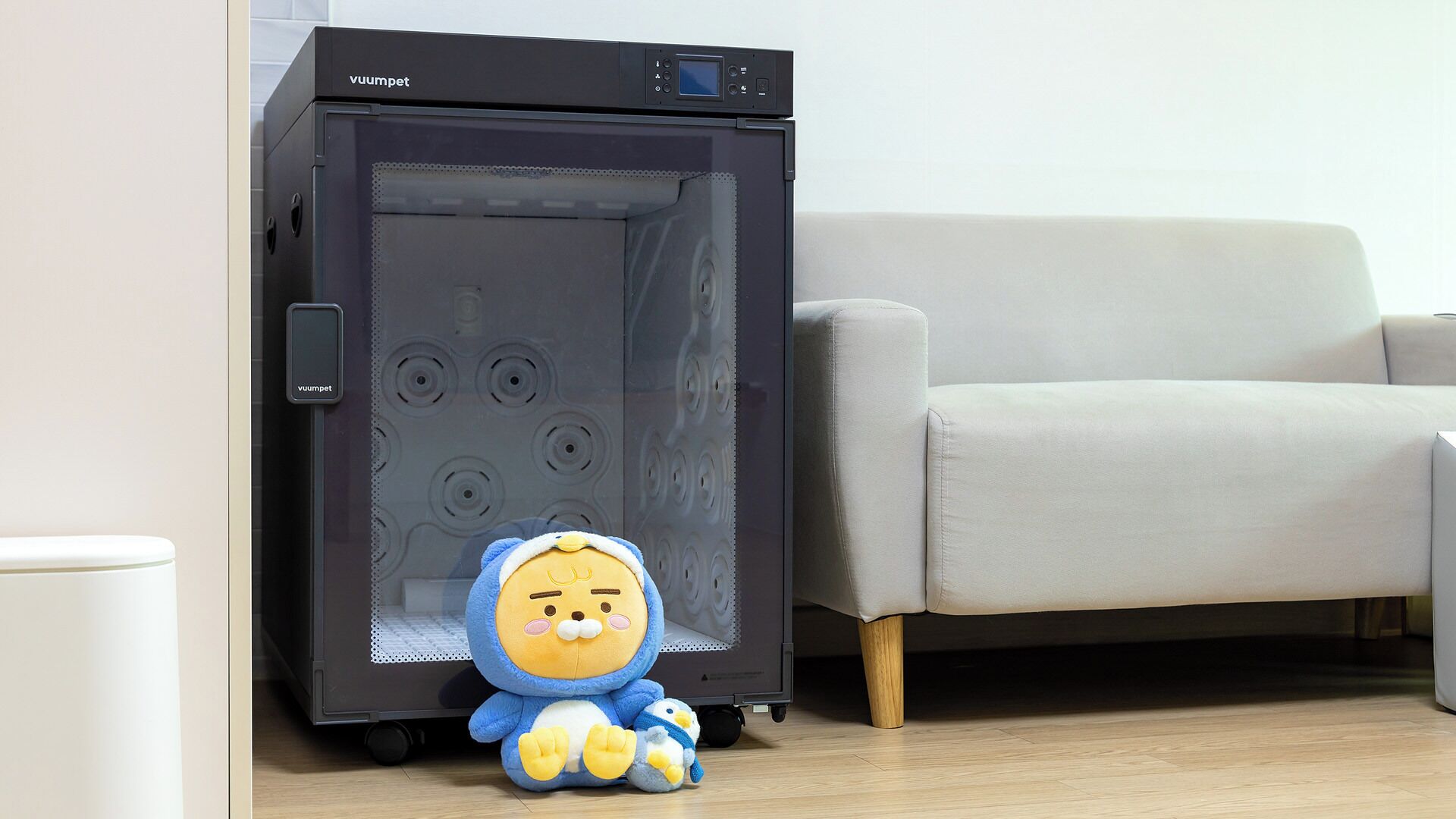 Ein Zimmer mit einem schwarzen Gerät, einem grauen Sofa und einem Kuscheltier.