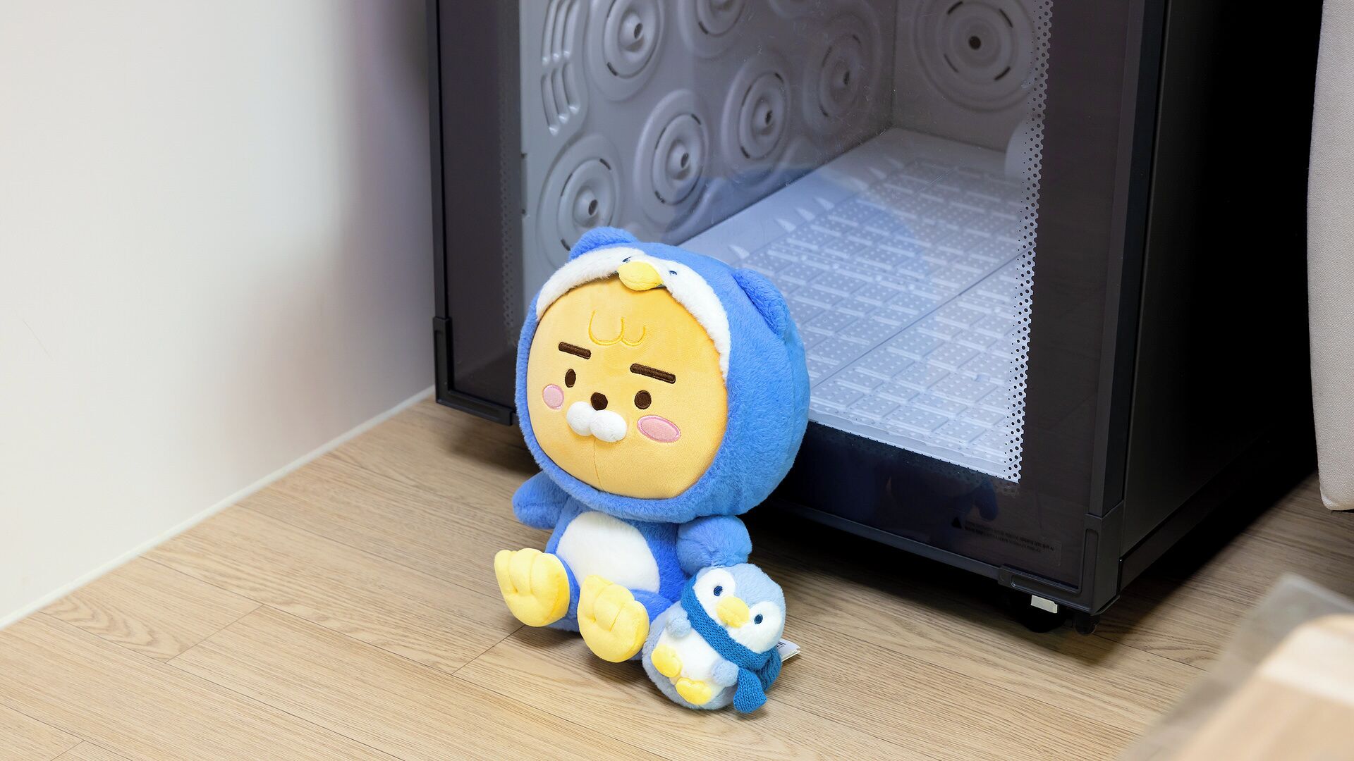 A plush toy in a blue outfit sitting on a wooden floor next to a microwave.