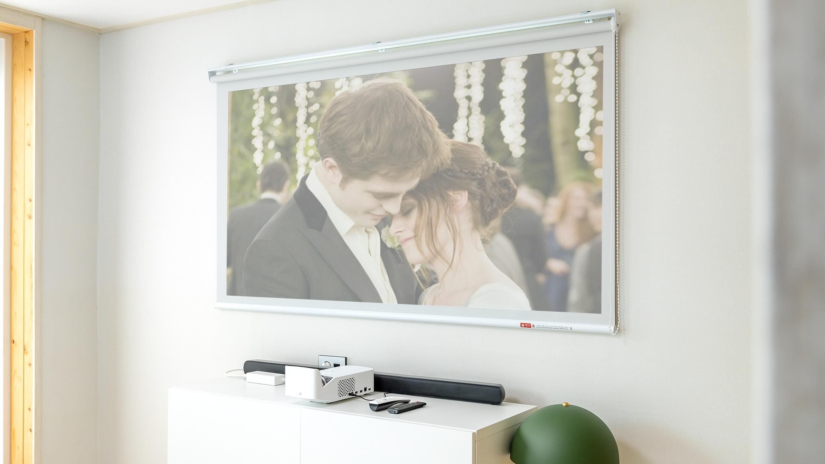 Una pantalla de proyector instalada en la pared muestra una foto de boda. Debajo de la pantalla, hay un mueble blanco con un altavoz, un control remoto y una esfera verde decorativa.