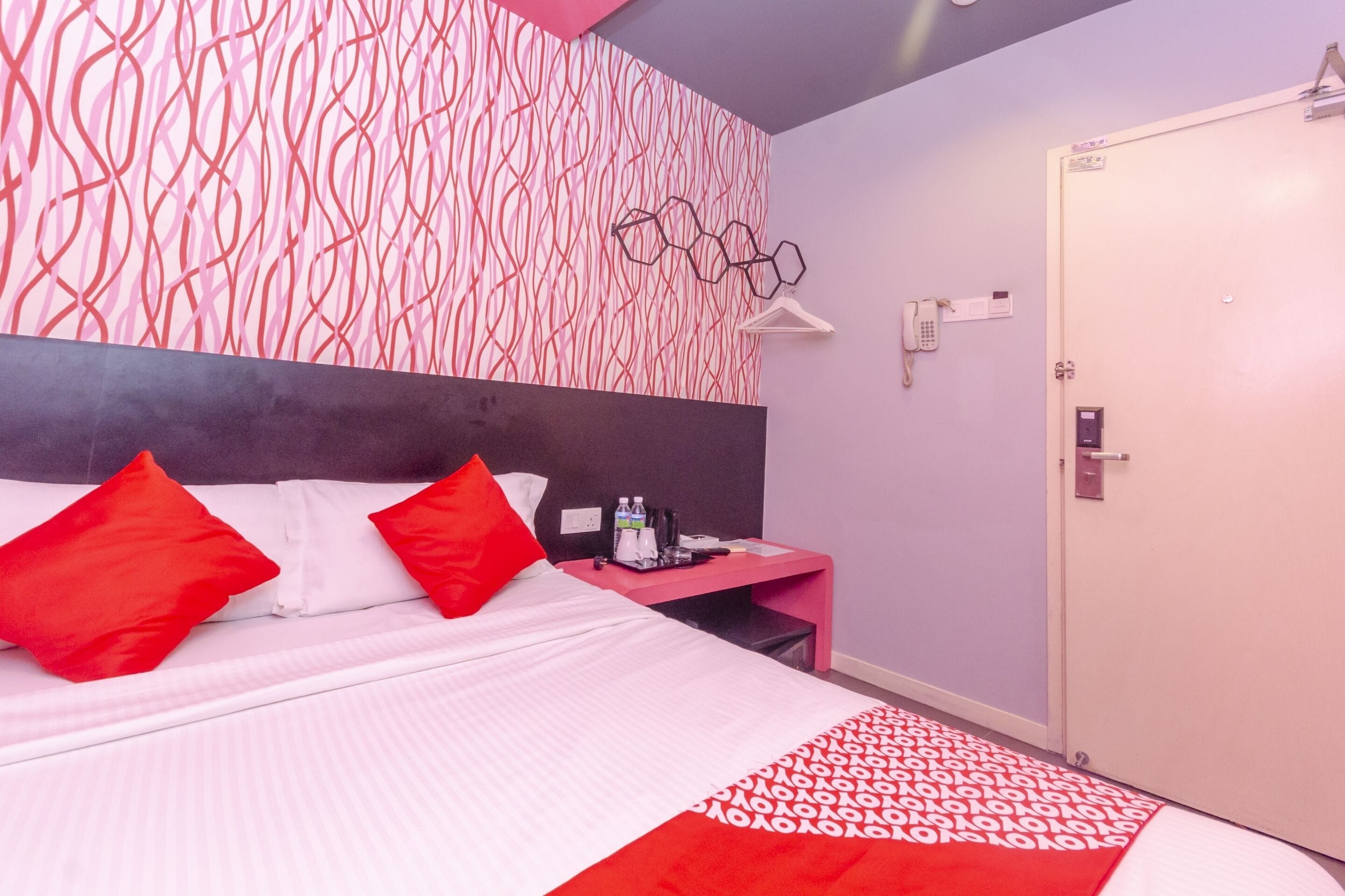 A modern hotel room with a large bed, red and white bedding, a pink nightstand, and a door with a lock.