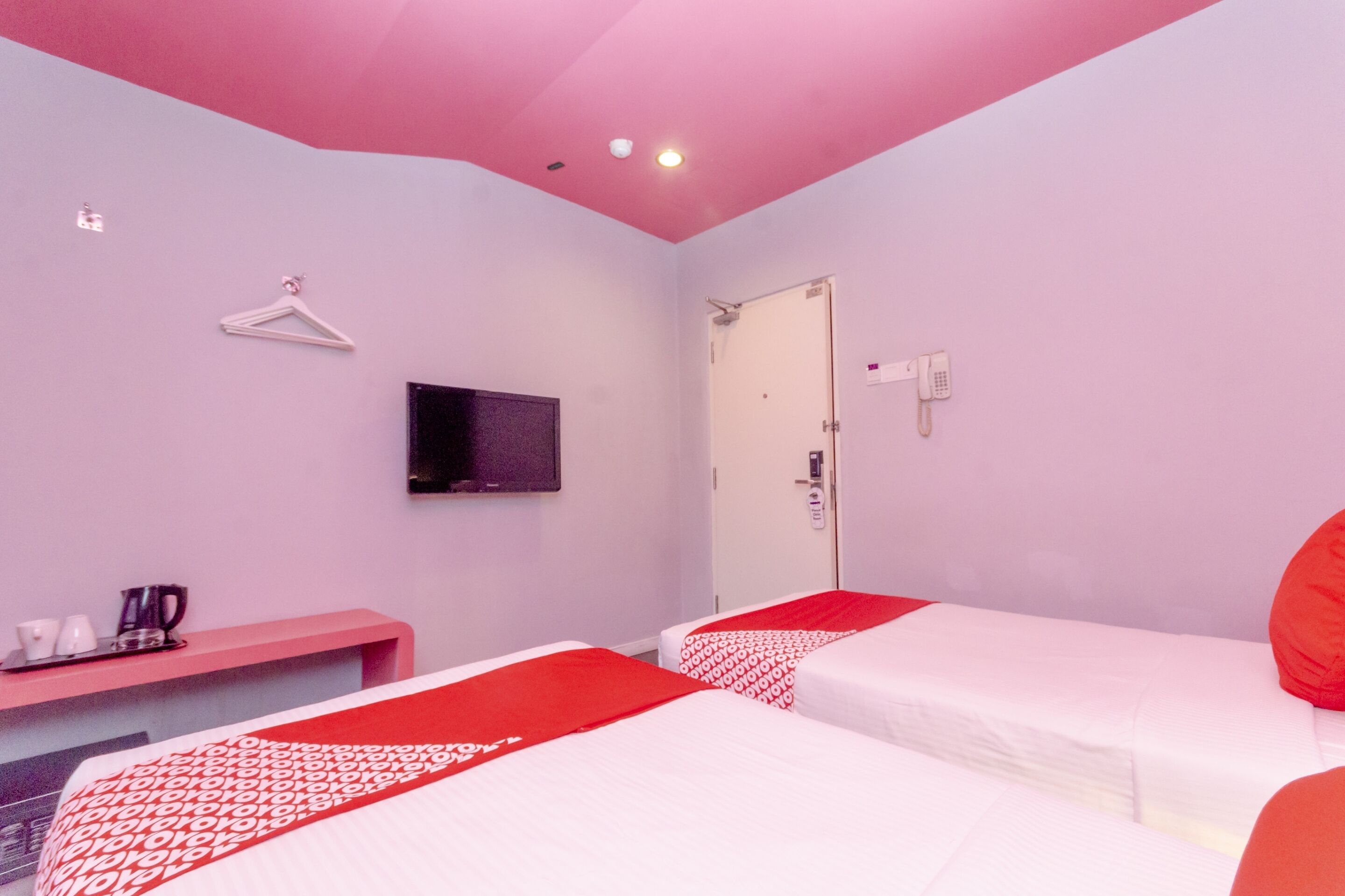A hotel room with two beds, a TV mounted on the wall, a desk with a kettle, cups, and a tray.