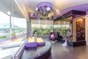 Un moderno vestíbulo de hotel con un área de estar curva, un sillón morado y un gran ventanal que ofrece vistas a zonas verdes y un puente.