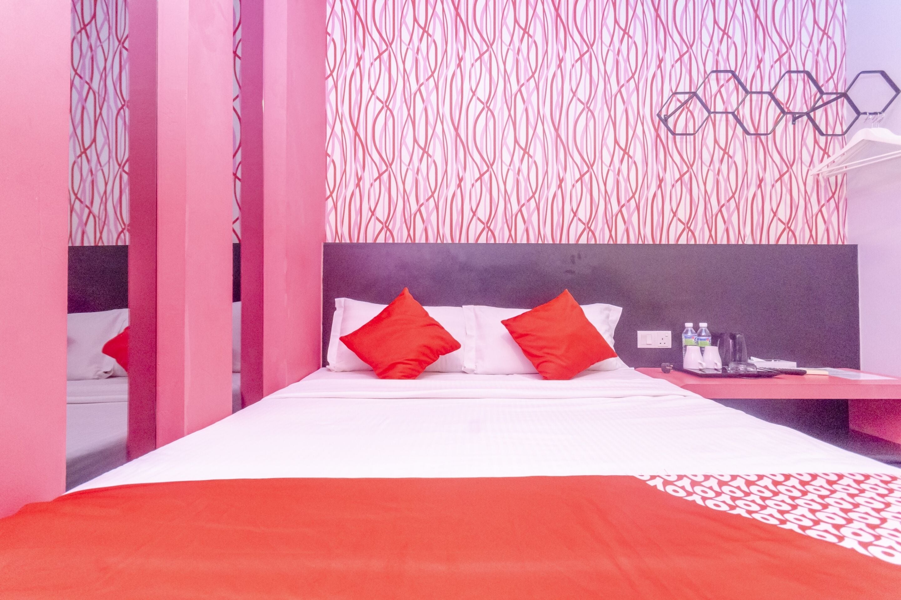 A modern hotel room with a large bed, red pillows, a nightstand with a lamp, and a patterned wall.