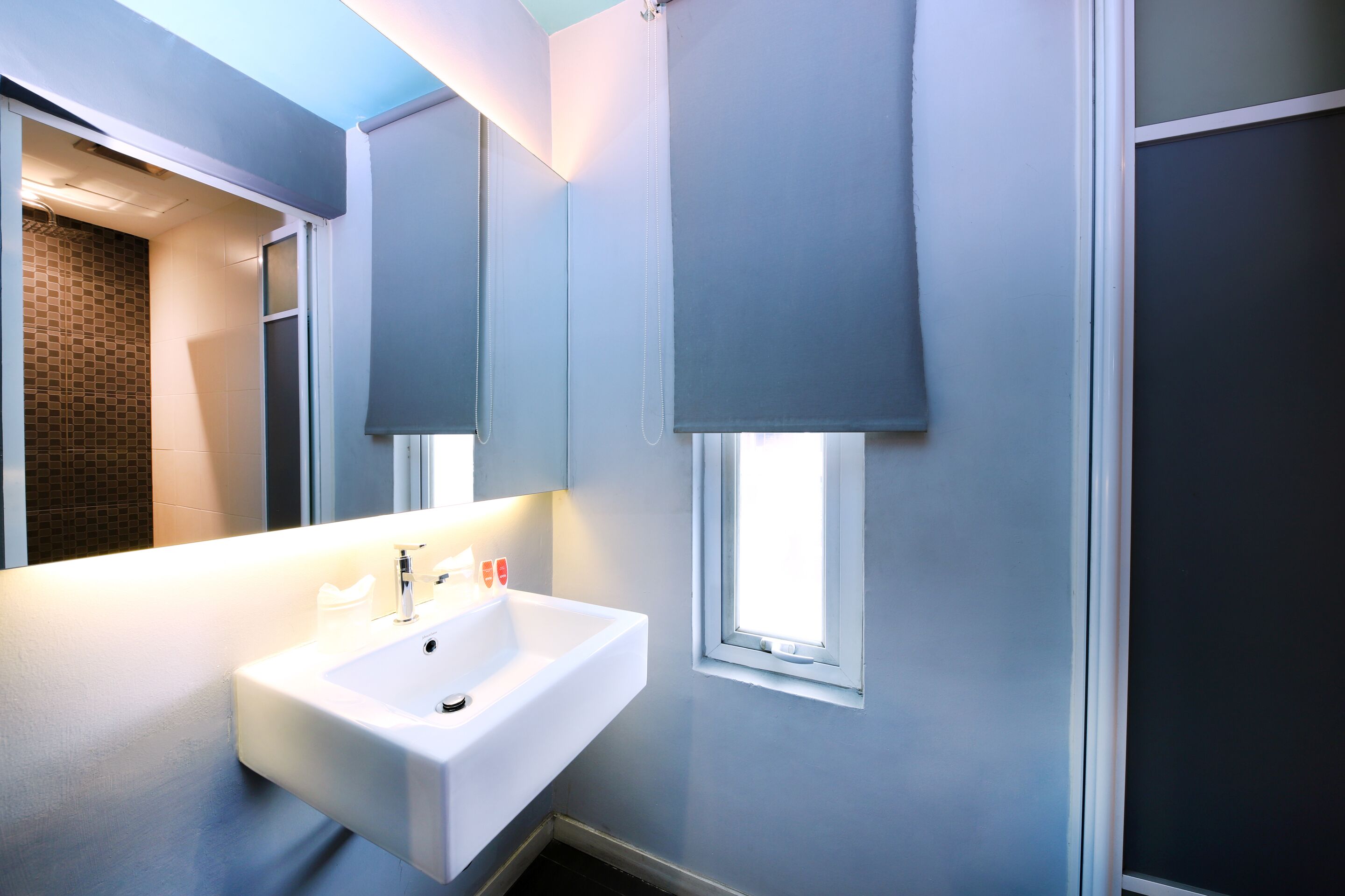 A modern bathroom with a white sink, a mirror, and a window with blinds.