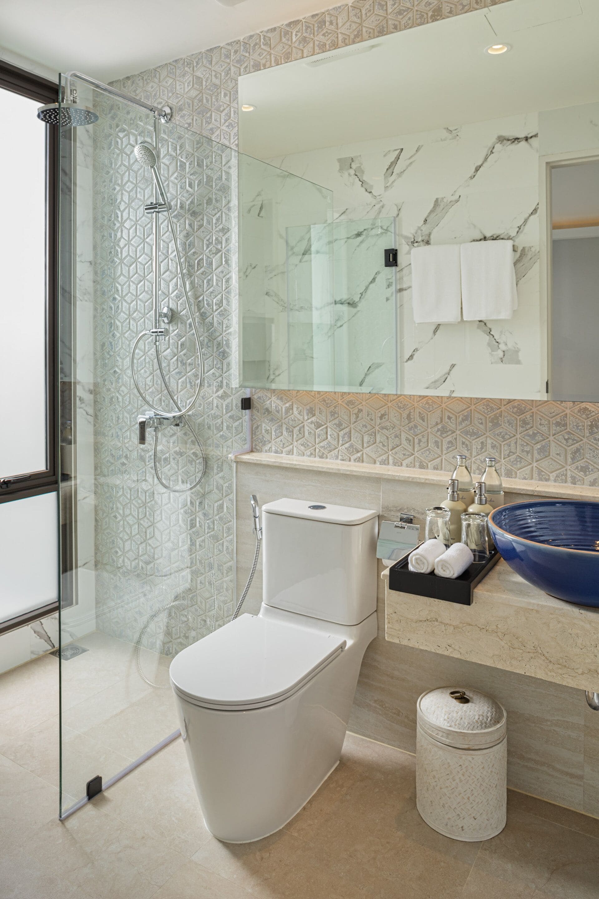 Un baño moderno con ducha de vidrio, inodoro blanco, pared con azulejos de mármol y un lavabo azul grande en el tocador.
