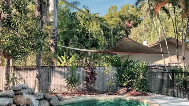 A swimming pool with a stone waterfall feature, surrounded by a wooden fence and various tropical plants.