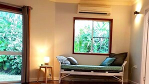 A room with a wooden floor, a bed with a green mattress and patterned pillows, a small table with a lamp, and a window with a view of greenery.