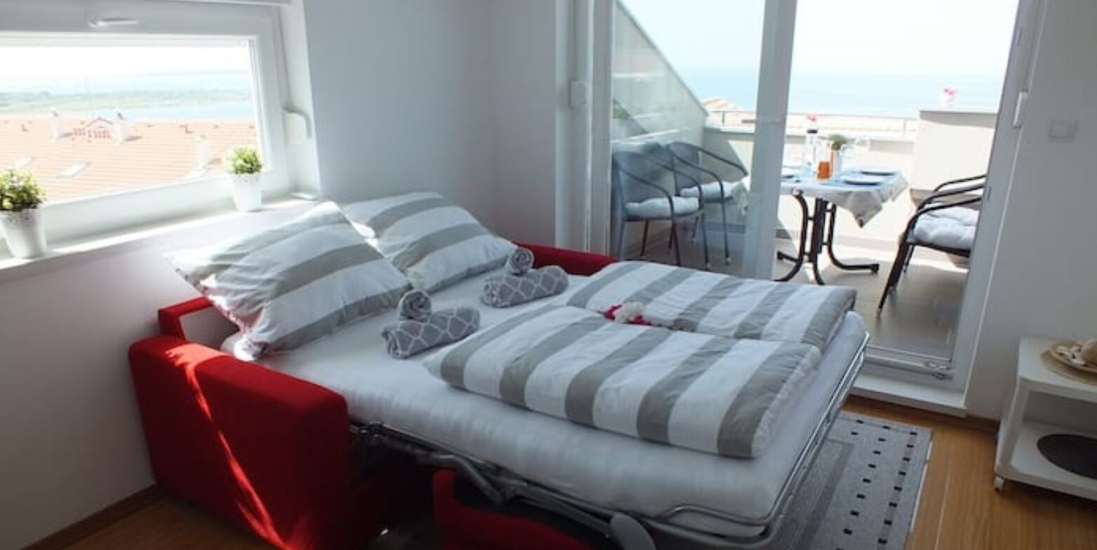 Ein kompaktes Bett mit gestreifter Bettwäsche, ein roter Stuhl und Blick auf den Strand durch ein großes Fenster.