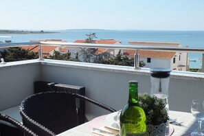 A balcony with a table set for two, a green bottle, and a glass. The view includes a body of water and buildings with red-tiled roofs. - Ferienwohnung "calimero" - mit Meerblick (Novalja)