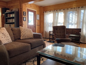 A cozy living room with a brown sofa, a glass coffee table, a wooden bookshelf, and a large window with curtains.